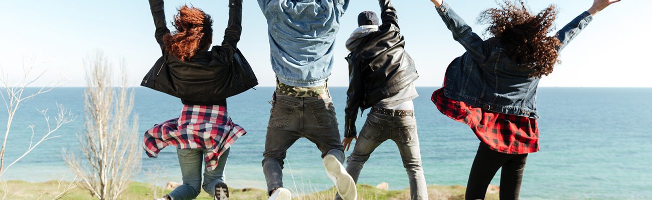 Back view picture of a group of friends jumping outdoors near beach with raised hands.